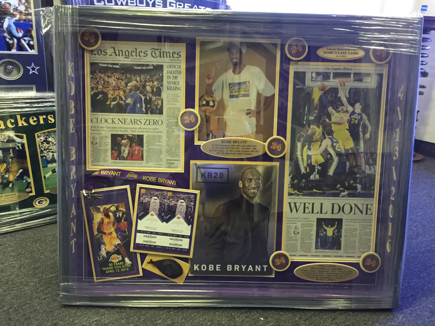 This collage is custom framed with components from Kobe Bryant’s final game in 2016. It includes a photo, confetti pieces, LA Times newspapers, commemorative tickets and more. It is a purple and yellow collage with a black frame.